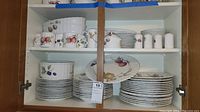 Photo of china dinner plates, salad plates, serving dishes, and mugs with fruit and nut motifs stored in a cabinet shelf.