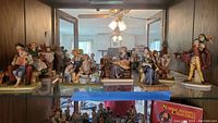 Shelf showing a group of Norman Rockwell figurines arranged in a lit glass cabinet, showcasing multiple figures in various poses and colors