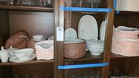 Wooden cabinet with multiple shelves holding white porcelain Rosenthal china including oval serving dishes, dinner plates, tea cups, and fabric storage bags in pink and brown.