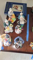 Six Royal Doulton figurines on wood surface with collector's book behind, showing detailed colorful characters.