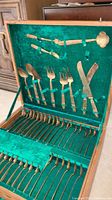 Full view of the opened wooden storage case showing various brass flatware pieces held in place by green velvet straps against a green velvet backing.