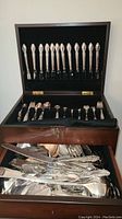 Open wooden case showing knives standing upright and other flatware in compartments below.
