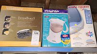 Three small kitchen appliances in original boxes: VillaWare InterBake 3 waffle maker, Cocomotion hot chocolate maker blender, George Foreman Colours grill.
