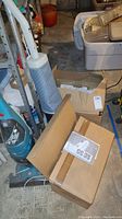 Photo showing the Hoover upright vacuum cleaner with blue/white body standing upright, alongside several cardboard boxes and a Floor Mate vacuum in a basement setting.
