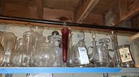 Wide view of all glassware items arranged on a shelf, showing decanters, vases, and pitchers in clear and cranberry glass.