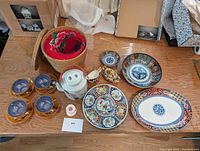 All items displayed on wooden table including the four teacups with saucers, teapot with floral design, woven basket with lid, and variety of decorative plates.