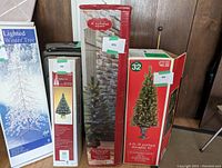 Pack of two boxed Christmas trees: one in a white box labeled 'Lighted Winter Tree,' another in a red box labeled '32 4 ft. lit potted Douglas fir,' a third box with St. Nicholas Square branding, tall and rectangular.