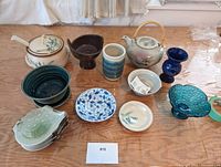Photo of collection of stoneware items on wood-grain table, showing teapot, plates, bowls, vases and ladle