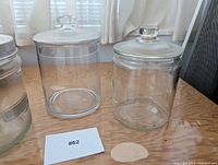 Front view of two cookie jars with lids placed on a wooden surface by a window with curtains.