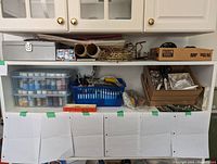 Wide view of two shelves filled with various craft materials including metal cash box, holiday cracker boxes, paint storage containers, brushes, rolled papers, and decorative brass frame.