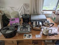 Full view of electric kitchen appliances on a table including George Foreman grill, Crock-Pot slow cooker, toaster, kettle, slow cookers, coffee maker box, oven mitts