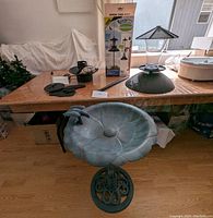 Full view of bird bath fully assembled on floor beneath a table showing decorative metal stand and basin with bird figurines. Parts for bird feeder or fountain on table above.