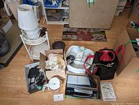 View of assorted household and office items on wooden floor showing lamp, coffee pot, kettle, steamer rack, electronic accessories, and box with papers