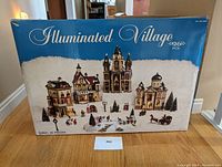 Front view of 24-inch long box showing full set of 26 illuminated Christmas village pieces including buildings, figurines, trees, and horse-drawn carriage.