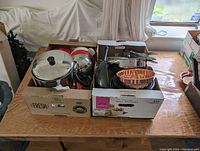 Photo of boxes on table containing kitchen cookware including a stainless steel stew pot lid visible, kettle, and copper wok with ribbed interior.