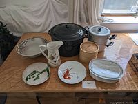 Top view photo showing all kitchen items on a wooden table including canning pot, metal pot, ceramic pitcher, wooden steamer basket, stainless steel oval dish, and three decorative plates.