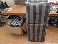 Lounger cushion packaged standing upright next to a box of six matching seat cushions still sealed in plastic.
