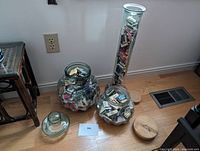 Three glass containers filled with matchbooks and matchboxes, displayed on a wooden floor. Two round jars with wooden lids are on the ground, and one tall glass cylindrical container is full and upright.