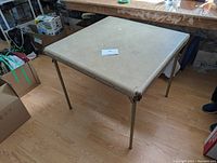 Square folding playing card table with beige vinyl surface and metal legs. Used condition with slight wear visible on edges.
