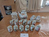 Full tea set including 12 cups, teapot with stand, sugar and creamer vessels with stand displayed on table with a clock and other items in background.