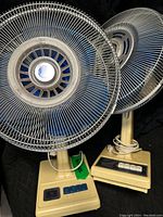 View of two cream-colored vintage electric fans side by side with blue blades and metal grilles. Each has multi-speed blue buttons on the base and cream pedestal stems.