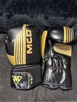Pair of black and gold MCD boxing gloves laid out on black textured surface, showing front and wrist strap with 20 oz size indicator.