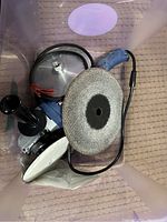 Electric grinder or buffer with abrasive disc, detachable handle, and metal guard inside a plastic bin.