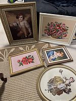 Overview photo of five framed needlepoint artworks on carpet floor, showing a range of floral and figurative designs.