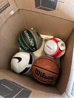 Photo showing five sports balls inside a box, including a Wilson basketball, a Spalding Extreme ball, and two soccer balls.