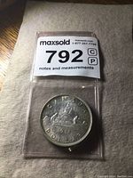Full view of the coin in protective packaging with MaxSold lot number.