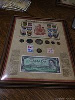 Full view of the framed Centennial collection showing all contents under glass including coins, stamps, and one dollar bill.