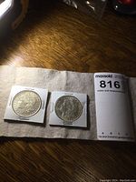 Both Morgan dollars in 2×2 cardboard holders displayed side by side on cloth with MaxSold notes and measurements card.