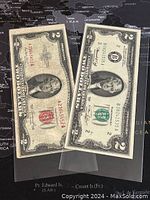 Two US $2 banknotes side by side, showing front designs and seals, displayed on a dark map background.