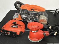 Image showing the three tools together on a black surface: a belt sander, a jigsaw, and a random orbital sander. All are Black & Decker branded and appear lightly used.