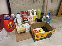 Photo shows entire lot of assorted cleaning and laundry supplies on a cardboard base against concrete wall. Items include Tide laundry detergent, Glad garbage bags box, Bull Dog steel soap, Lysol cleaner, various spray bottles, dish soap, laundry soap box, dryer sheets, several brushes, and broom.