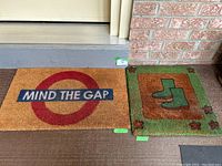 Photo showing two coir door mats side by side on front porch. Left: rectangular 'Mind The Gap' mat with red circle and blue text. Right: square green mat with boot and leaf design.