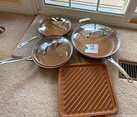 Set of three Gotham Steel fry pans with glass lids and one Gotham Steel grill pan all placed on carpeted floor.