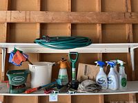 Wide shot of hose and garden accessories on wall shelf including garden hose coiled, hand seed spreader, bucket, hand sprayer, garden ties, spray bottles, garden gloves, and tools.