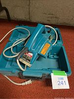 Makita power planer inside its blue carrying case, showing the tool plugged into power cord and lying against open case.