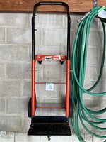 Full frontal view of metal dolly showing orange frame, black handle, and black platform plate with wheels.