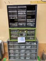 Three stacked plastic drawer organizers in black, green, and gray containing assorted screws and hardware items.