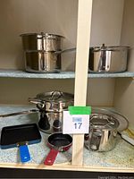 Image shows stainless steel pots and double boilers of varying sizes with lids, plus two small non-stick pans with colorful handles on a shelf.