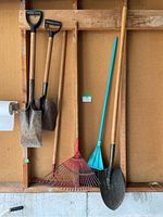 Full view of five garden tools hanging on a wooden wall in a garage: three shovels and two rakes of different designs and colors.