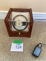 Stauer watch winder display case with wristwatch inside and charging cord beside it on carpeted floor