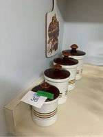 Four vintage metal kitchen canisters with lids arranged in descending size, cream colored with brown lids and decorative horizontal stripes.
