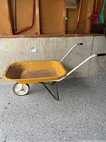 Yellow metal wheelbarrow with white handles, black grips, and single front wheel on a gray garage floor with tools hanging on wall.