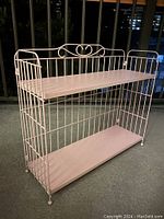 Full view of pink folding metal shelf photographed indoors on a carpeted floor under artificial light, showing two shelves and the metal frame.