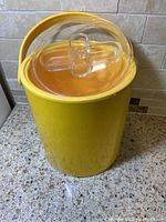 Front view of tall yellow cylindrical ice bucket with clear amber lid and yellow handle, on a kitchen countertop.
