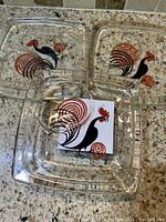 Three square glass snack plates with black and red/orange stylized rooster design, arranged on a granite counter.