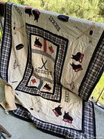 Quilt blanket displayed hanging outside showing central hockey-themed design with maple leaf, skates, hockey sticks, and puck patterns, bordered by plaid fabric.
